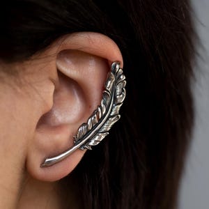 May include: A silver-colored ear cuff in the shape of a feather. The feather design is detailed with intricate lines and textures, giving it a realistic appearance. The cuff curves around the ear, creating a unique and stylish accessory.