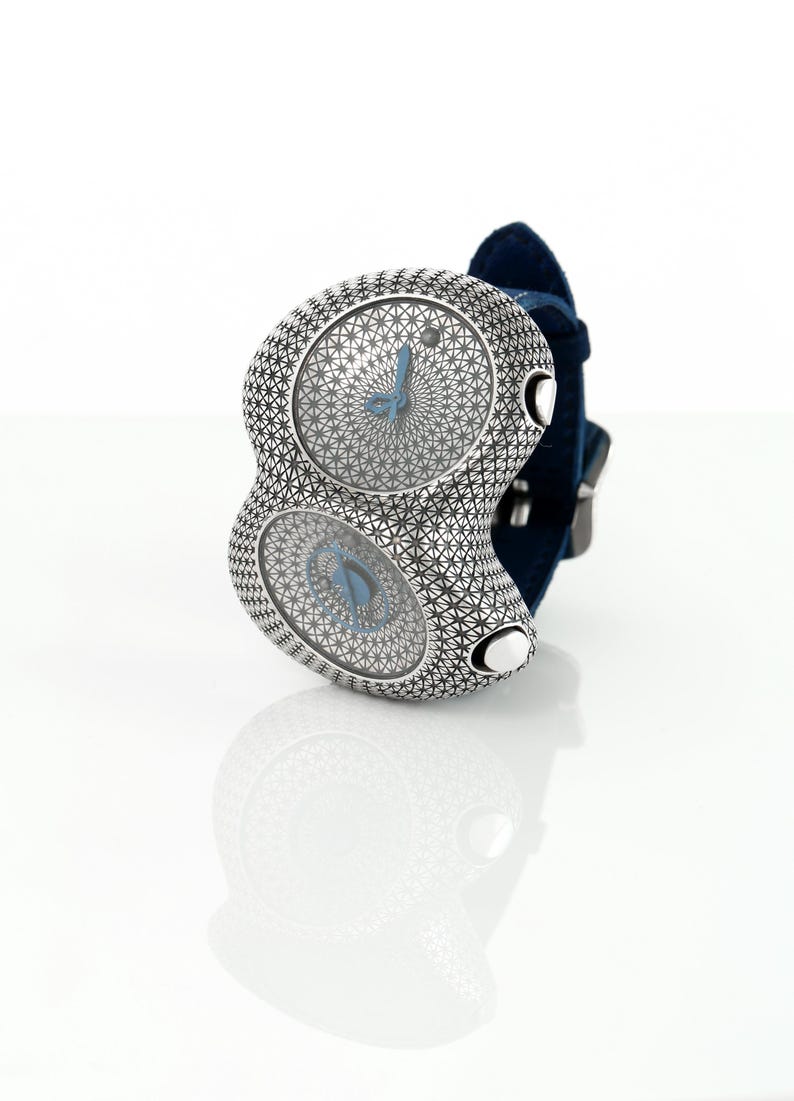 May include: A unique silver-toned wristwatch with two circular faces, each featuring a blue clock face and hands. The watch has a patterned design and is attached to a dark blue fabric band. The watch is displayed on a white surface.