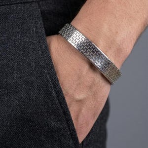 May include: A silver-toned metal cuff bracelet featuring a repeating geometric pattern of interlocking L-shaped lines. The bracelet has a wide, flat band design with a polished finish that reflects light across its textured surface.