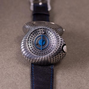 May include: A silver-toned wristwatch with a geometric pattern on the face and band. The watch face has a blue center with blue hands. The black leather band has blue stitching. The watch is on a neutral background.