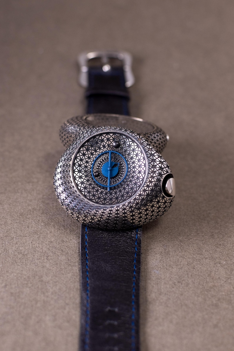May include: A silver-toned wristwatch with a geometric pattern on the face and band. The watch face has a blue center with blue hands. The black leather band has blue stitching. The watch is on a neutral background.