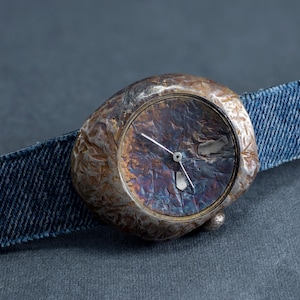 May include: A unique wristwatch with a textured, metallic face and a denim band. The watch face has a dark, iridescent finish with silver hands. The denim band is a classic blue color, creating a contrast with the watch face.