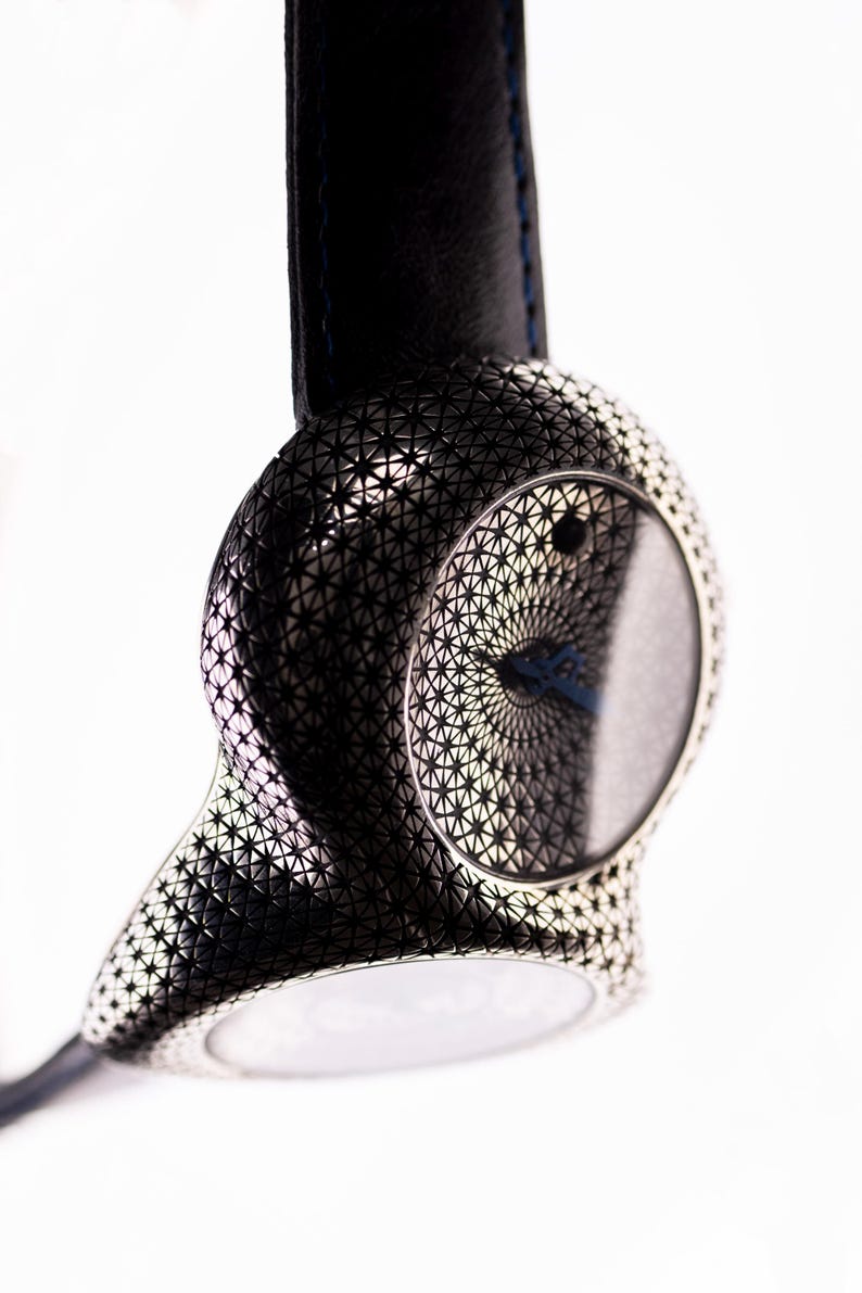 May include: A modern wristwatch with a black leather band and a silver-toned case. The case features a geometric pattern and a circular watch face with a blue hand. The watch is suspended against a white background.