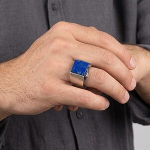 May include: A silver-toned ring featuring a large, rectangular, deep blue stone set horizontally on the band. The ring has a wide, smooth metal shank that tapers slightly toward the palm, showcasing a bold, geometric design with a polished finish.