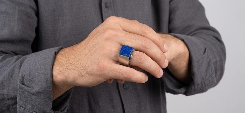 May include: A silver-toned ring featuring a large, rectangular, deep blue stone set horizontally on the band. The ring has a wide, smooth metal shank that tapers slightly toward the palm, showcasing a bold, geometric design with a polished finish.