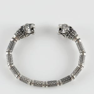 Puede incluir: Brazalete plateado con terminales de cabeza de león detalladas. La banda del brazalete tiene una textura tejida con acentos cilíndricos segmentados. Las cabezas de león están intrincadamente esculpidas, mostrando bocas abiertas y melenas detalladas. Fondo blanco.