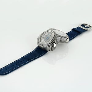 May include: A unique wristwatch with a dual-dial design. The watch features a silver-toned case with a detailed pattern and a navy blue suede strap. The watch has a silver buckle. The watch is a luxury item.