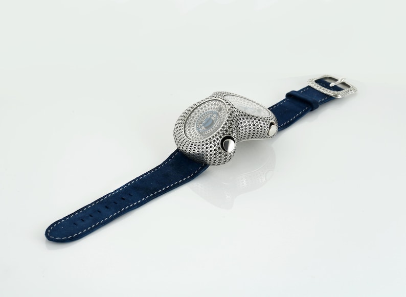 May include: A unique wristwatch with a dual-dial design. The watch features a silver-toned case with a detailed pattern and a navy blue suede strap. The watch has a silver buckle. The watch is a luxury item.