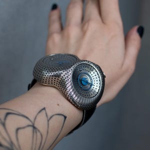 May include: A unique silver-toned wristwatch with a patterned design and a black leather band. The watch features two circular faces with blue accents. The watch is worn on a wrist with a floral tattoo.
