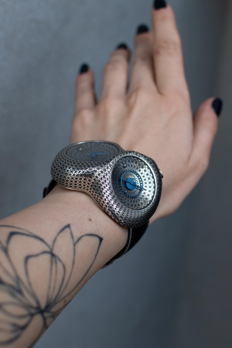 May include: A unique silver-toned wristwatch with a patterned design and a black leather band. The watch features two circular faces with blue accents. The watch is worn on a wrist with a floral tattoo.