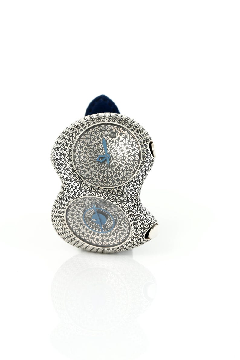 May include: A unique silver-toned wristwatch with two circular watch faces. The watch features a geometric pattern and blue hands. A dark blue band is attached to the top of the watch. The watch is displayed on a white surface.