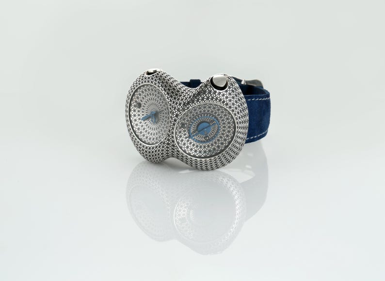 May include: A unique silver wristwatch with two circular faces, each featuring a detailed starburst pattern and blue hands. The watch has a dark blue suede strap. The watch is displayed on a reflective surface.