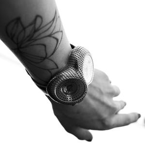 May include: A black and white image of a unique wristwatch. The watch features a detailed, patterned design and is worn on a wrist with a floral tattoo. The watch's face is visible, and the overall aesthetic is modern and artistic.