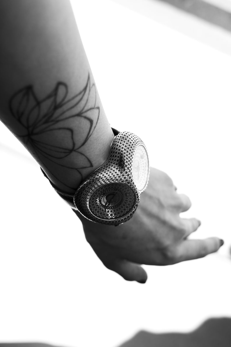 May include: A black and white image of a unique wristwatch. The watch features a detailed, patterned design and is worn on a wrist with a floral tattoo. The watch's face is visible, and the overall aesthetic is modern and artistic.