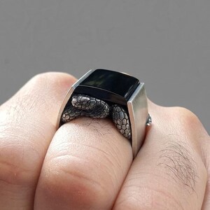 May include: A silver ring featuring a rectangular black stone set in a smooth metal band. The sides of the ring are decorated with a detailed, textured snake motif that wraps around the base of the stone setting, creating a distinct contrast.