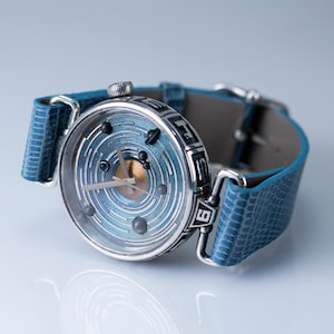 Pode incluir: Um relógio de pulso único com um mostrador azul com um design em espiral e pequenos detalhes escuros. O relógio tem uma caixa prateada e uma pulseira de couro azul. O mostrador do relógio tem cerca de 3,8 cm de diâmetro.