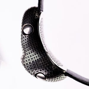 May include: Close-up of a modern wristwatch with a black leather strap. The watch case is dark gray with a geometric pattern and two silver buttons. The watch is set against a white background.