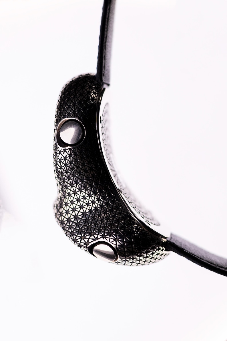 May include: Close-up of a modern wristwatch with a black leather strap. The watch case is dark gray with a geometric pattern and two silver buttons. The watch is set against a white background.