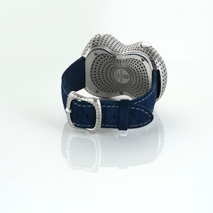 May include: A luxury wristwatch with a silver-toned, textured face and a dark blue suede strap. The watch features a decorative buckle with a diamond-like design. The watch is displayed on a white surface.