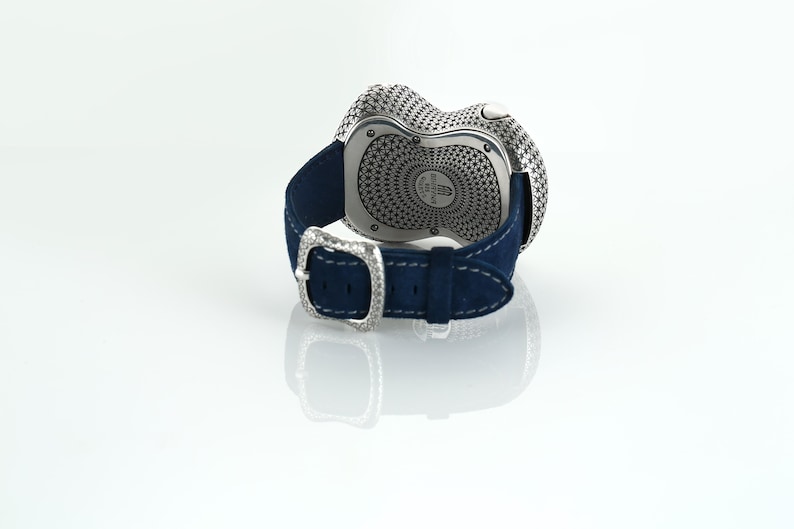May include: A luxury wristwatch with a silver-toned, textured face and a dark blue suede strap. The watch features a decorative buckle with a diamond-like design. The watch is displayed on a white surface.