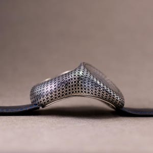 May include: A silver-toned ring with a detailed geometric pattern, resting on a dark blue leather strap. The ring's design features a repeating star-like motif, creating a textured surface. The background is a neutral, muted brown.