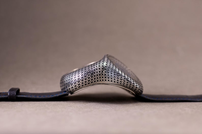 May include: A silver-toned ring with a detailed geometric pattern, resting on a dark blue leather strap. The ring's design features a repeating star-like motif, creating a textured surface. The background is a neutral, muted brown.