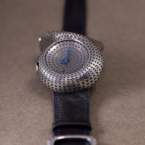 May include: A silver-toned wristwatch with a patterned face and a blue hand. The watch has a black leather band with blue stitching and a silver buckle. The watch face has a geometric design.