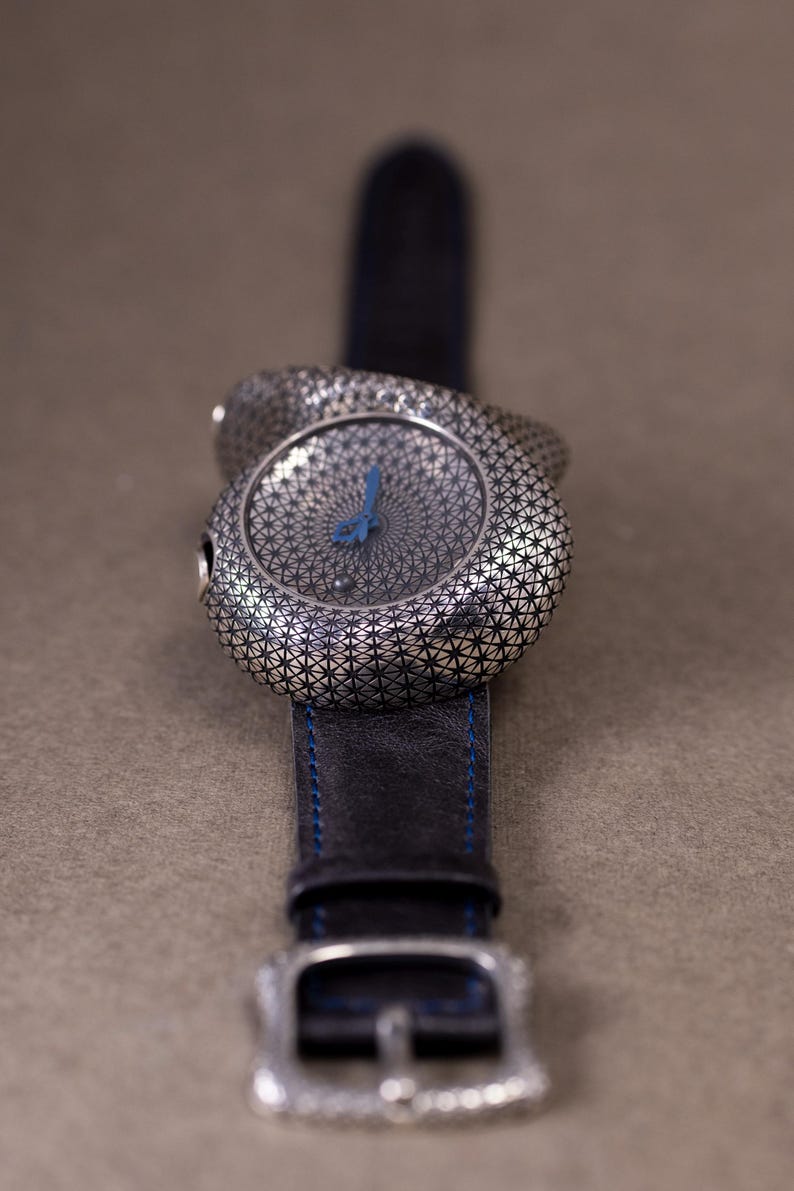 May include: A silver-toned wristwatch with a patterned face and a blue hand. The watch has a black leather band with blue stitching and a silver buckle. The watch face has a geometric design.