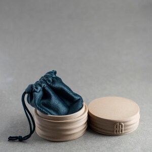 May include: A round, light beige ceramic container with a ribbed exterior and a matching lid featuring a small embossed logo. A dark teal velvet drawstring pouch is tucked inside the open container, with its dark teal cord hanging over the side.