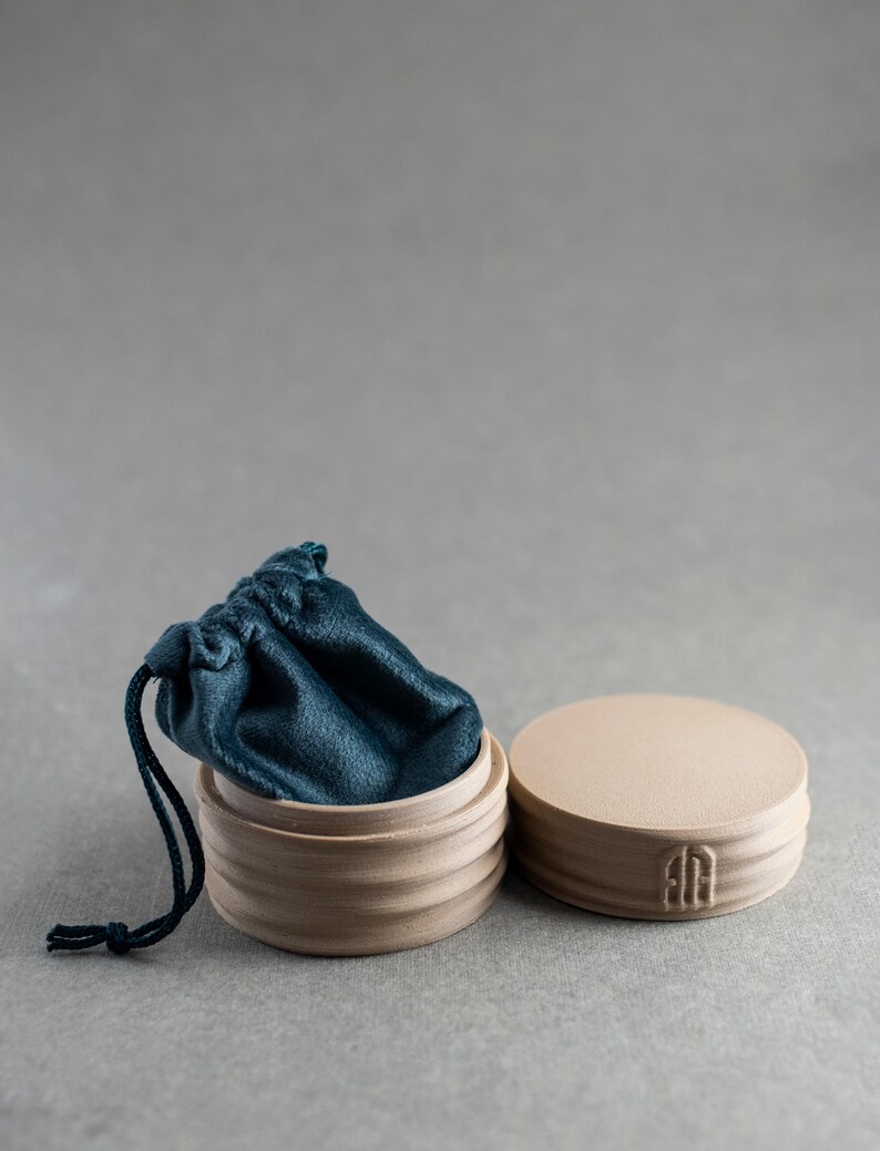 May include: A round, light beige ceramic container with a ribbed exterior and a matching lid featuring a small embossed logo. A dark teal velvet drawstring pouch is tucked inside the open container, with its dark teal cord hanging over the side.
