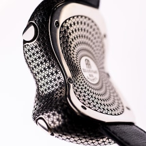 May include: Close-up of a luxury wristwatch with a black leather strap. The watch case is silver-toned with intricate geometric patterns. The watch face features a circular design and the brand name. The watch is set against a white background.