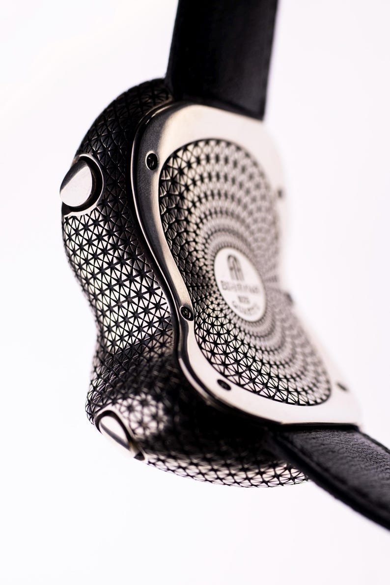 May include: Close-up of a luxury wristwatch with a black leather strap. The watch case is silver-toned with intricate geometric patterns. The watch face features a circular design and the brand name. The watch is set against a white background.