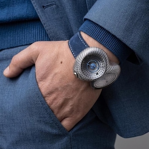 May include: A luxury wristwatch with a unique, textured silver-tone case and a blue dial. The watch is worn on a blue suede strap. The person is wearing a blue sweater and a blue suit jacket. The watch is a statement piece.