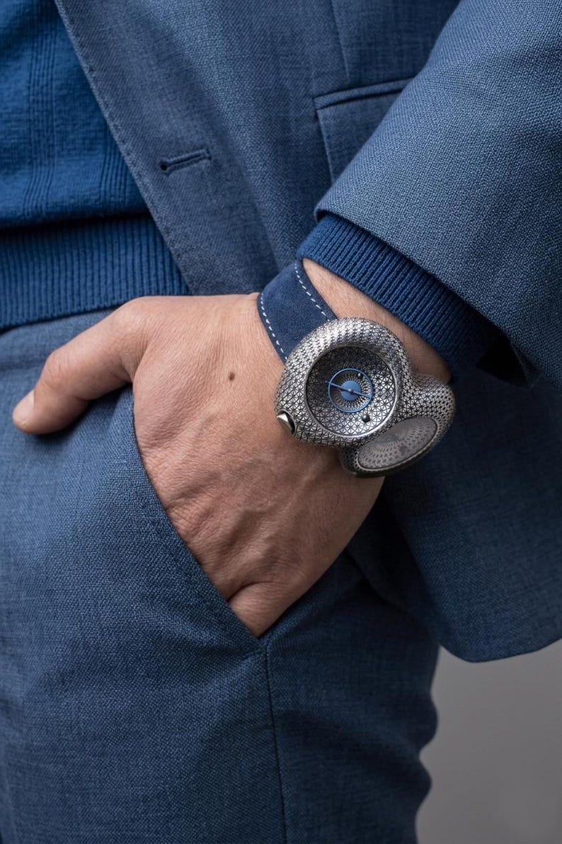 May include: A luxury wristwatch with a unique, textured silver-tone case and a blue dial. The watch is worn on a blue suede strap. The person is wearing a blue sweater and a blue suit jacket. The watch is a statement piece.