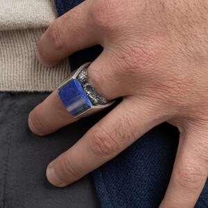 May include: A silver ring featuring a rectangular, deep blue lapis lazuli stone set into a band with a textured, scale-like pattern on the sides. The ring has a polished metal finish and a wide, substantial band design that wraps around the finger.