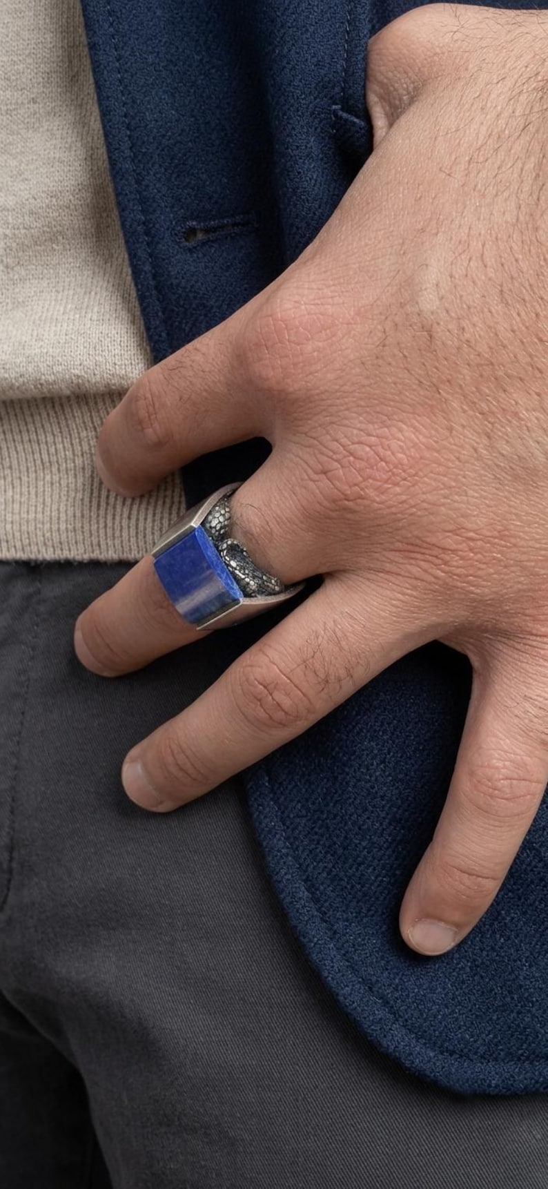 May include: A silver ring featuring a rectangular, deep blue lapis lazuli stone set into a band with a textured, scale-like pattern on the sides. The ring has a polished metal finish and a wide, substantial band design that wraps around the finger.