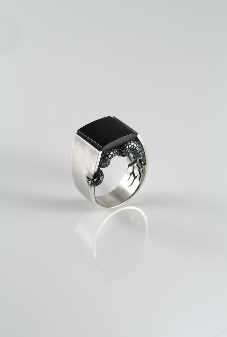 May include: A silver ring with a large, square black onyx stone. The ring features a detailed, textured design on the sides. The ring is displayed on a white surface, showcasing its reflective qualities. A modern piece of jewelry.