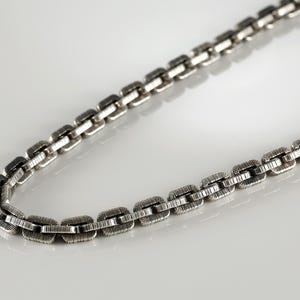 Könnte beinhalten: Eine silberfarbene Halskette mit einem strukturierten, rechteckigen Kettenglied-Design. Die Halskette wird auf einer weißen Oberfläche präsentiert, wobei die Glieder das Licht reflektieren. Die Kette ist gebogen.
