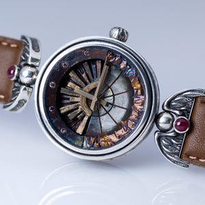 May include: A close-up of a unique wristwatch with a brown leather band. The watch face features a complex, multi-colored design with silver accents and a small, decorative crown. The band is attached with ornate silver clasps, each with a small, red gemstone.