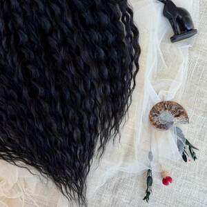 May include: A mass of long, wavy black hair extensions positioned next to a small black cat figurine, a spiral ammonite fossil, and a delicate sprig of dried greenery with a single small red flower bud on a textured off-white fabric background.