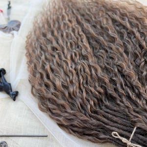 May include: A bundle of synthetic hair extensions featuring a wavy, crimped texture. The hair transitions from a dark brown shade at the base to a lighter, warm brown tone at the ends. The bundle is secured at the bottom with a thin, light-colored string.