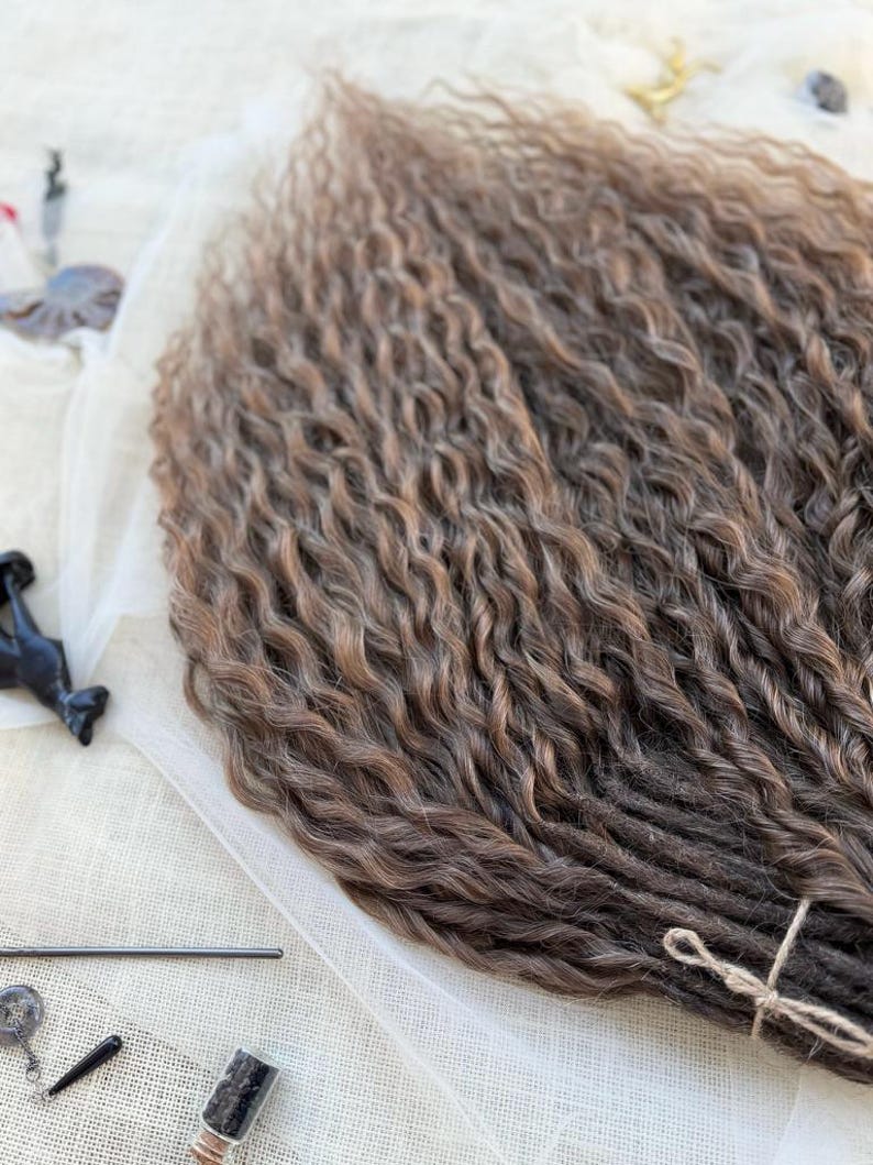 May include: A bundle of synthetic hair extensions featuring a wavy, crimped texture. The hair transitions from a dark brown shade at the base to a lighter, warm brown tone at the ends. The bundle is secured at the bottom with a thin, light-colored string.