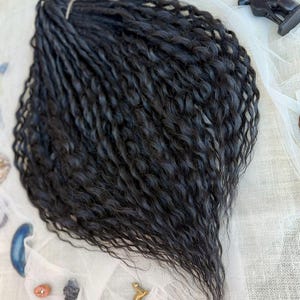 May include: A bundle of long, black, wavy hair extensions featuring a crimped texture. The strands are gathered at the top with a small band, creating a fan-like shape that tapers toward the bottom, showcasing a consistent dark color throughout.