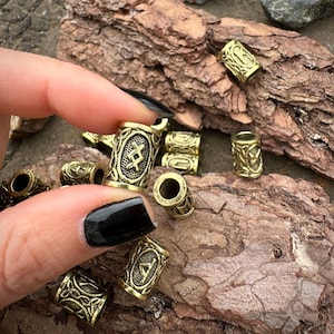 Op de afbeelding: Een verzameling antiek goudkleurige metalen kralen. De cilindrische kralen hebben gegraveerde patronen en symbolen, waaronder runen. De kralen worden getoond op een stuk getextureerd hout, waarbij één kraal door een hand wordt vastgehouden.