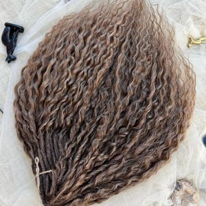 May include: A bundle of long, wavy, light brown synthetic hair extensions with a highlighted texture. The strands are gathered at one end and secured with a thin, light-colored string, creating a fan-like arrangement of individual wavy locks.