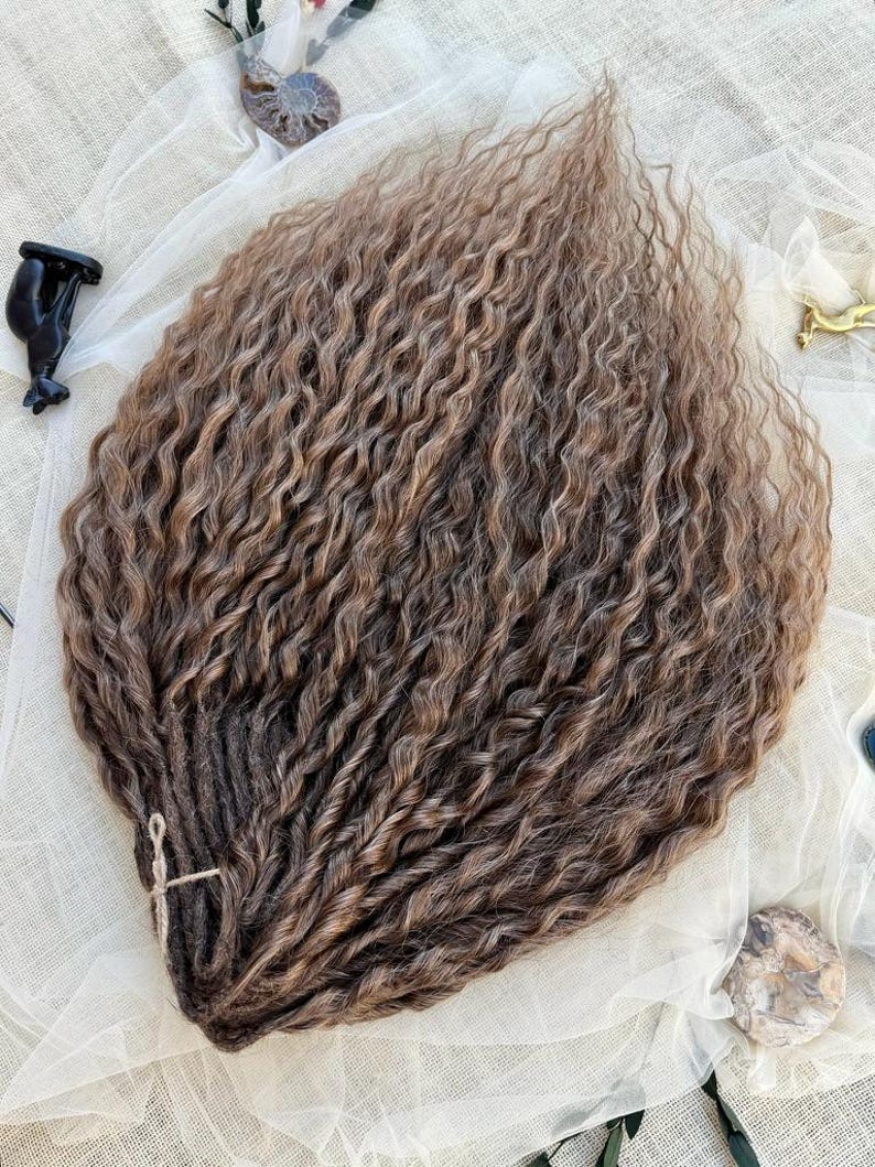 May include: A bundle of long, wavy, light brown synthetic hair extensions with a highlighted texture. The strands are gathered at one end and secured with a thin, light-colored string, creating a fan-like arrangement of individual wavy locks.