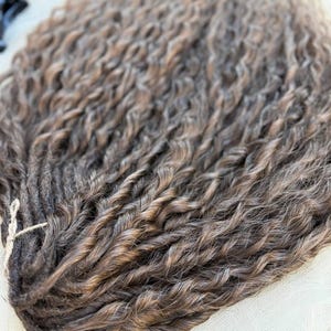 May include: A bundle of synthetic hair extensions featuring a wavy, textured pattern in a blend of dark brown and light brown highlights. The individual strands are twisted together and secured at the top with a thin, light-colored string.