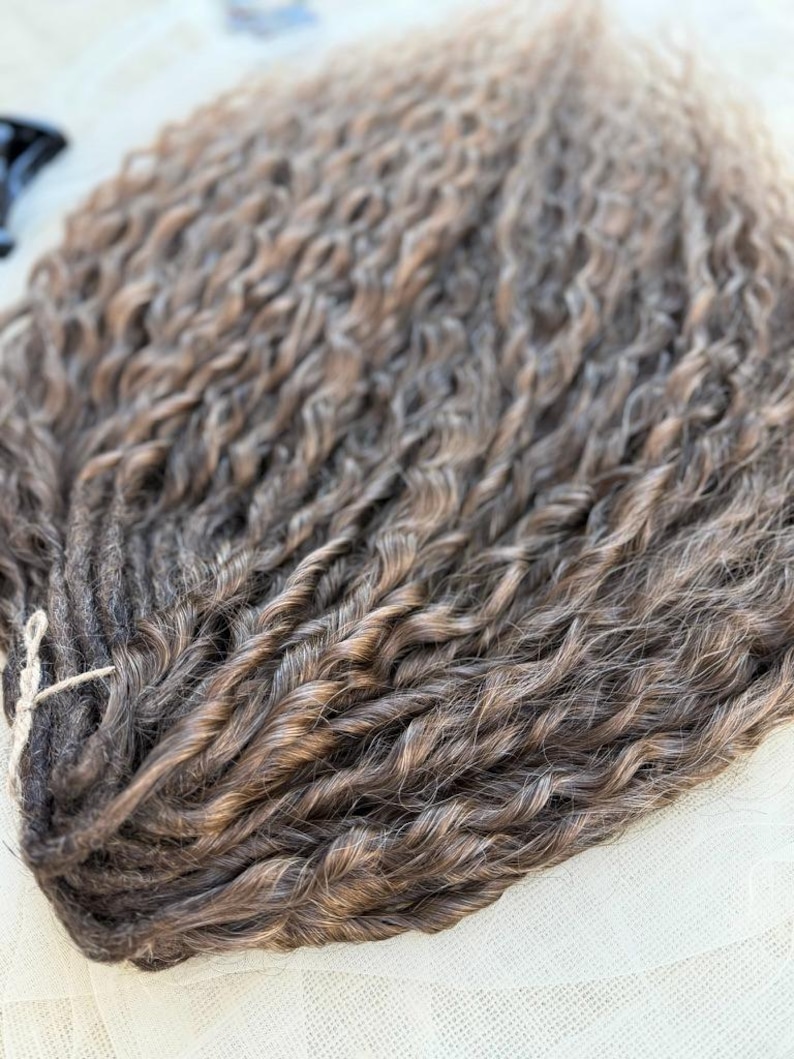 May include: A bundle of synthetic hair extensions featuring a wavy, textured pattern in a blend of dark brown and light brown highlights. The individual strands are twisted together and secured at the top with a thin, light-colored string.