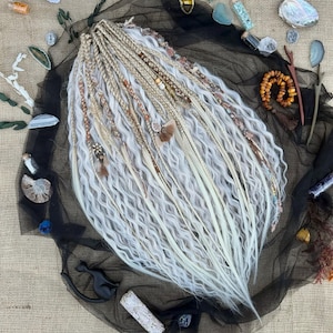 Puede incluir: Conjunto de rastas hechas a mano en tonos rubios y blancos, adornadas con cuentas y plumas. Las rastas están dispuestas sobre una tela de malla negra, rodeadas de cristales, conchas y otros elementos naturales. La estética general es bohemia y artística.