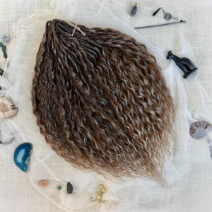 May include: A bundle of brown and blonde synthetic hair extensions with a wavy, twisted texture. The hair is arranged on a white fabric surface surrounded by small decorative items including a black cat figurine, stones, and a metal hair accessory.